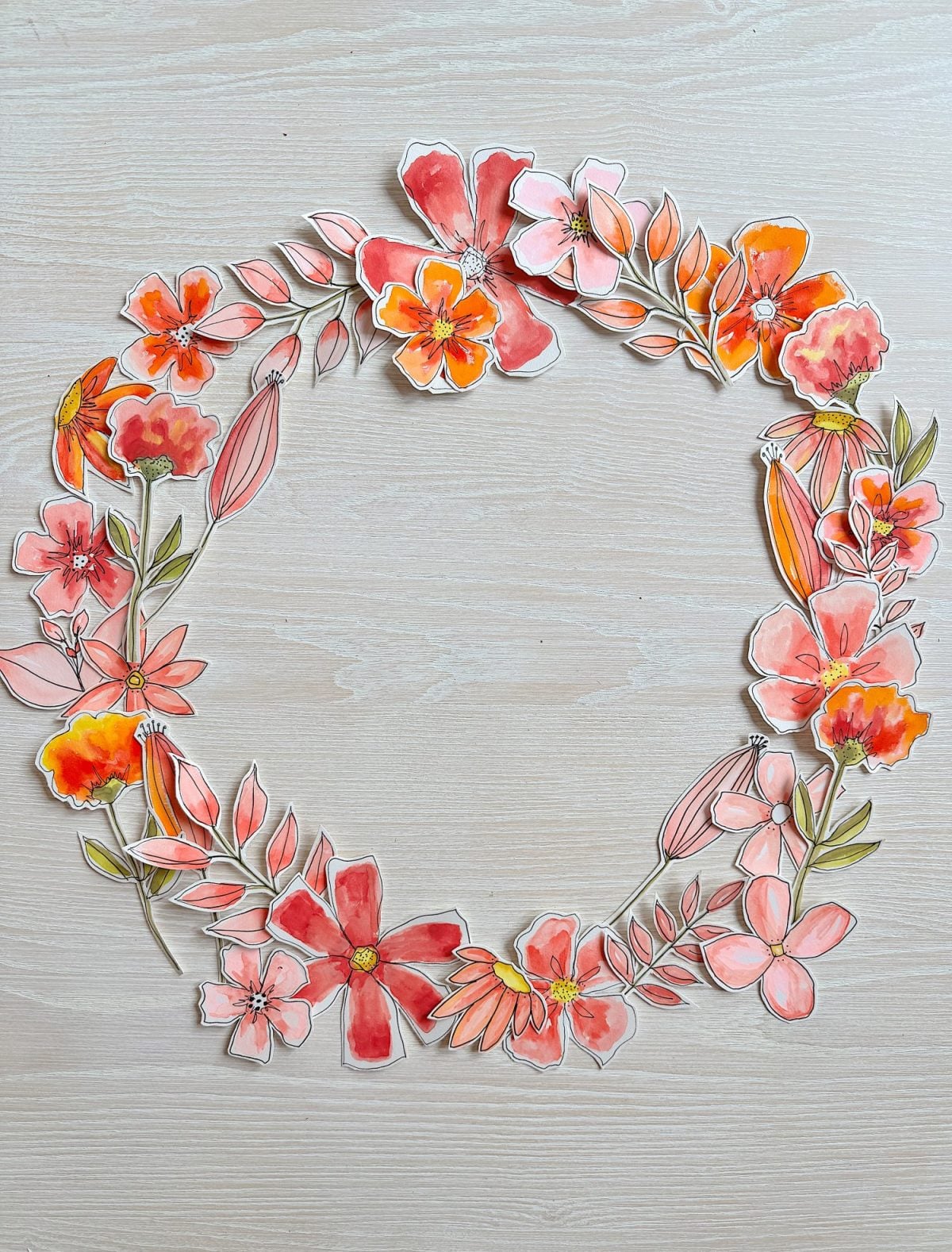 A circular floral wreath composed of hand-painted, cut-out orange, red, and pink flowers and green leaves arranged on a light wooden surface.
