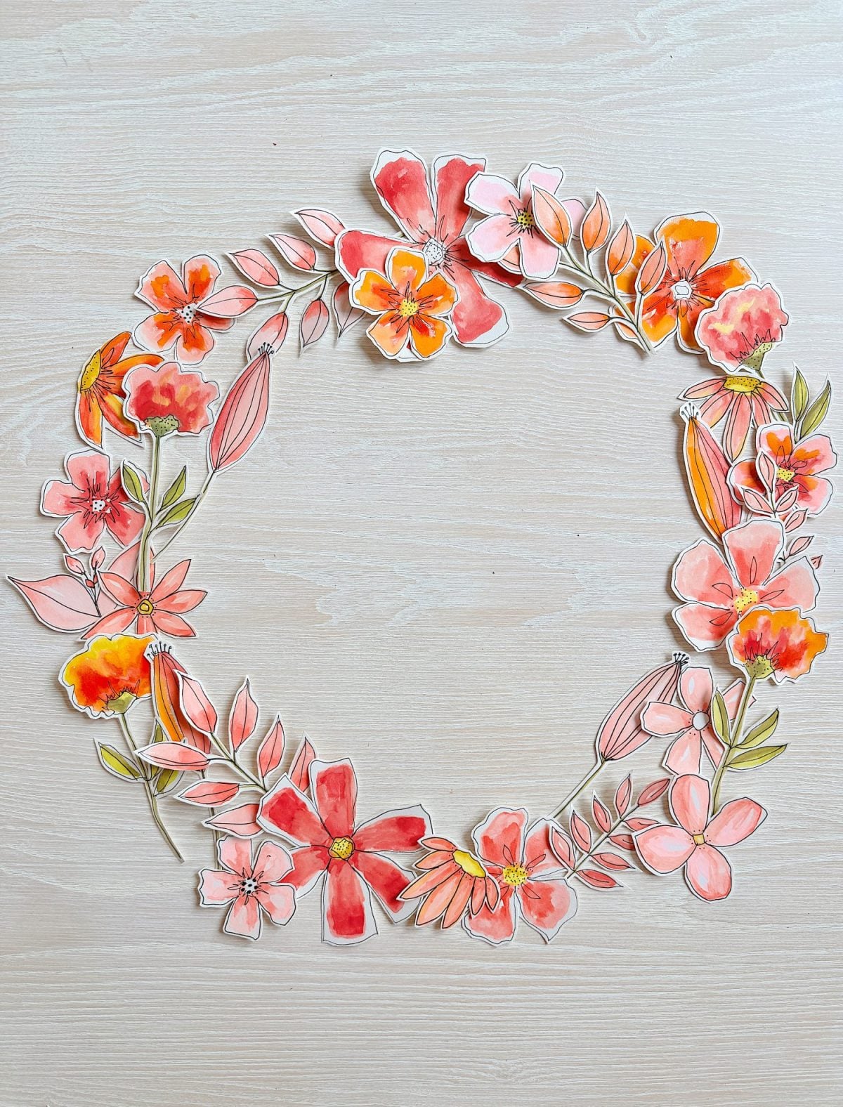 A handmade floral wreath with pink, orange, and red watercolor flowers and green leaves arranged in a circular pattern on a light wooden surface.