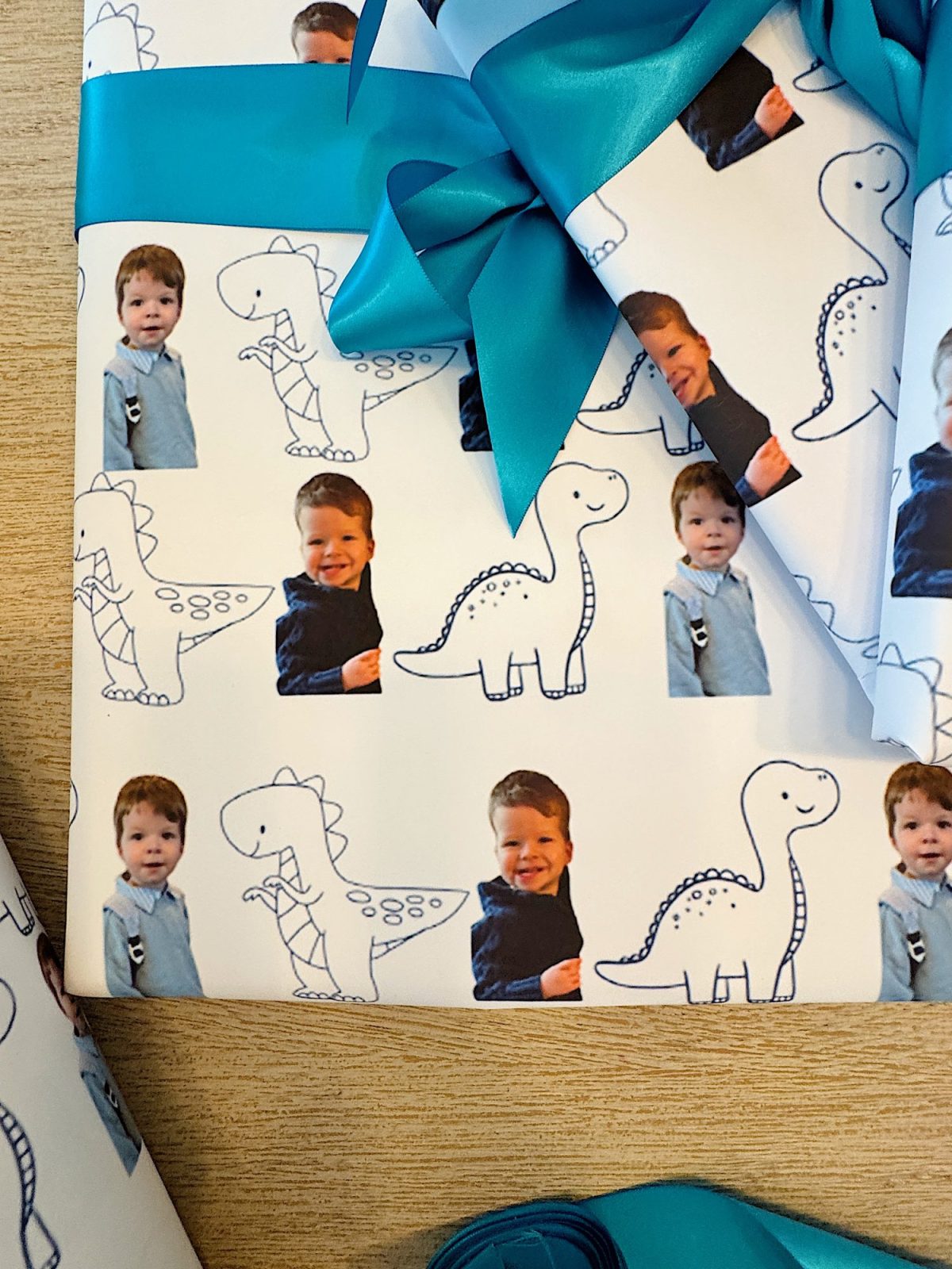 Gift wrapped in paper featuring alternating images of a young child and outlined dinosaur drawings, with a teal ribbon and bow on a wooden surface.