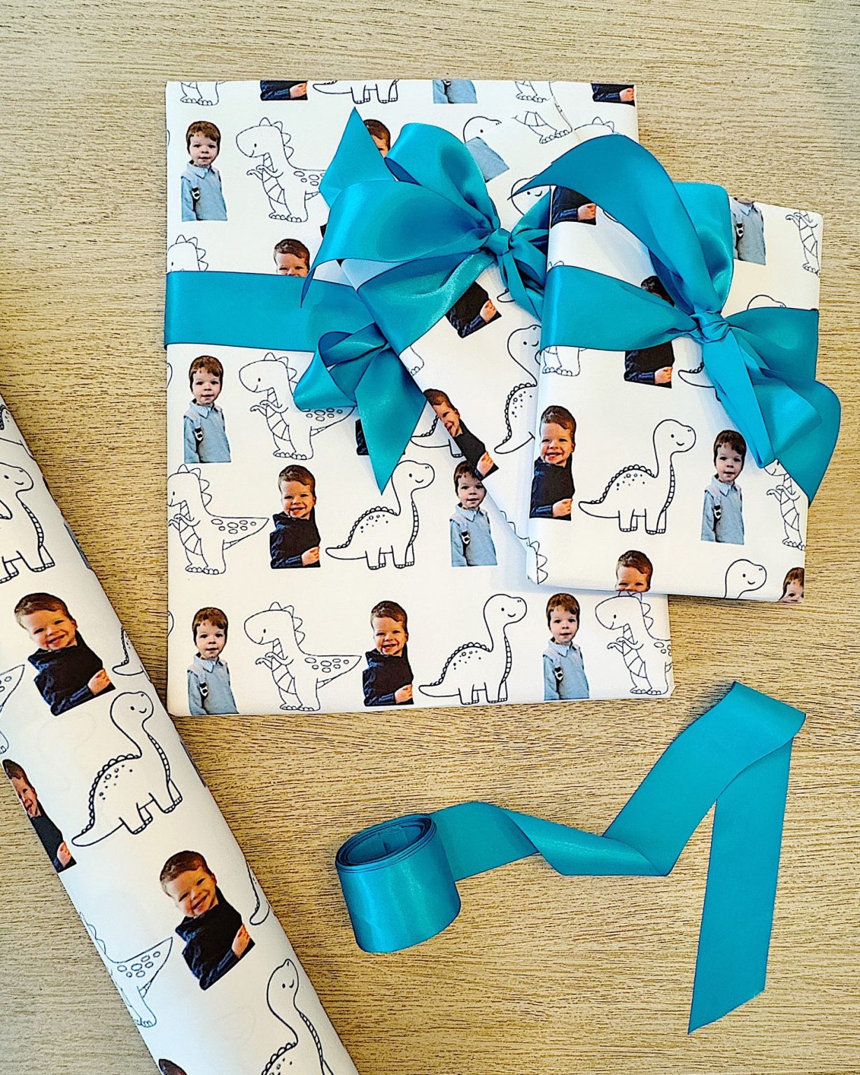 Gift boxes wrapped in white paper with dinosaur and child photo patterns, topped with blue ribbons. A roll of the same paper and a blue ribbon are nearby on a wooden surface.