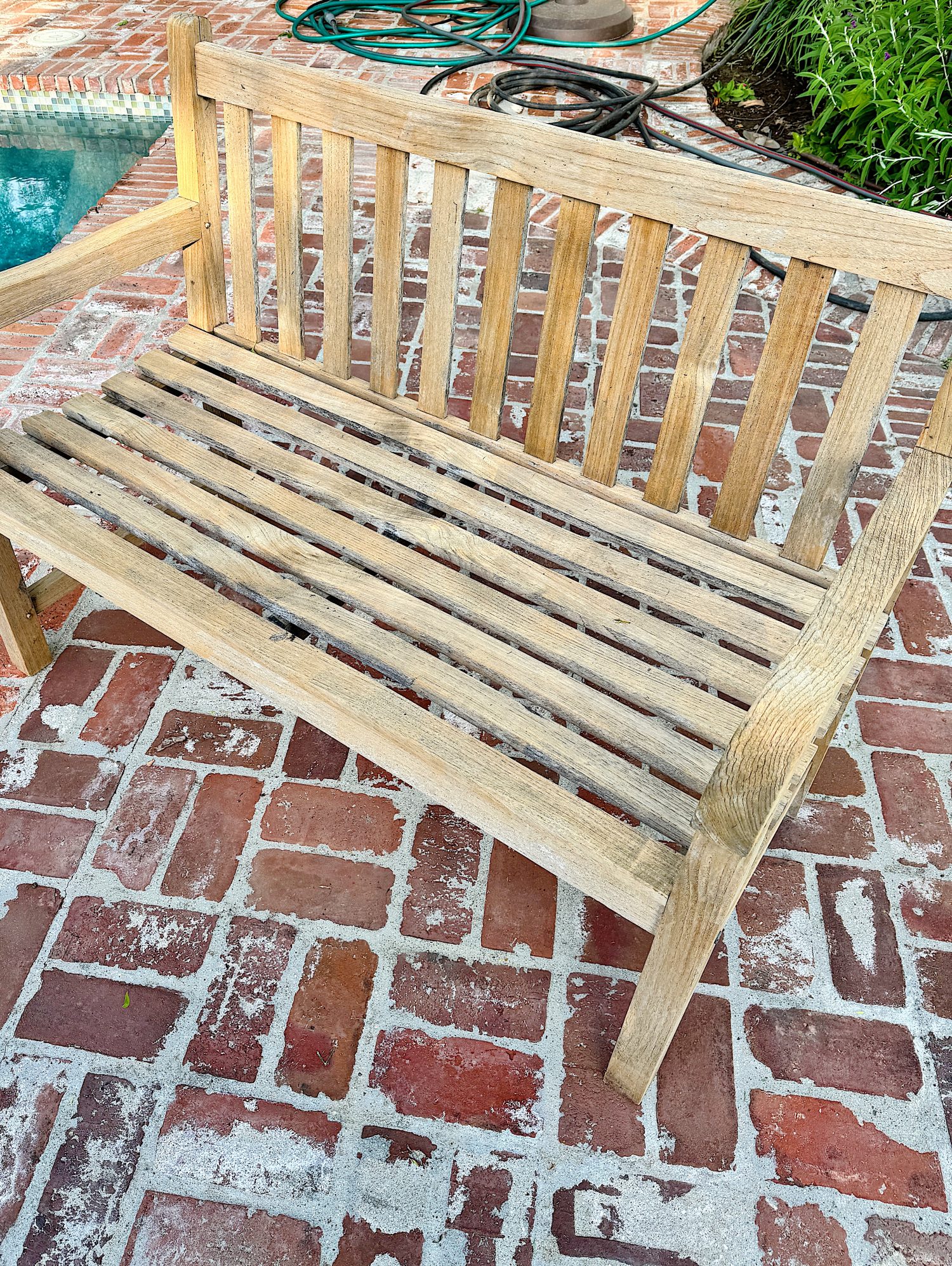 A wooden slatted bench sits on a red brick patio near the edge of a swimming pool, with a garden hose coiled in the background.