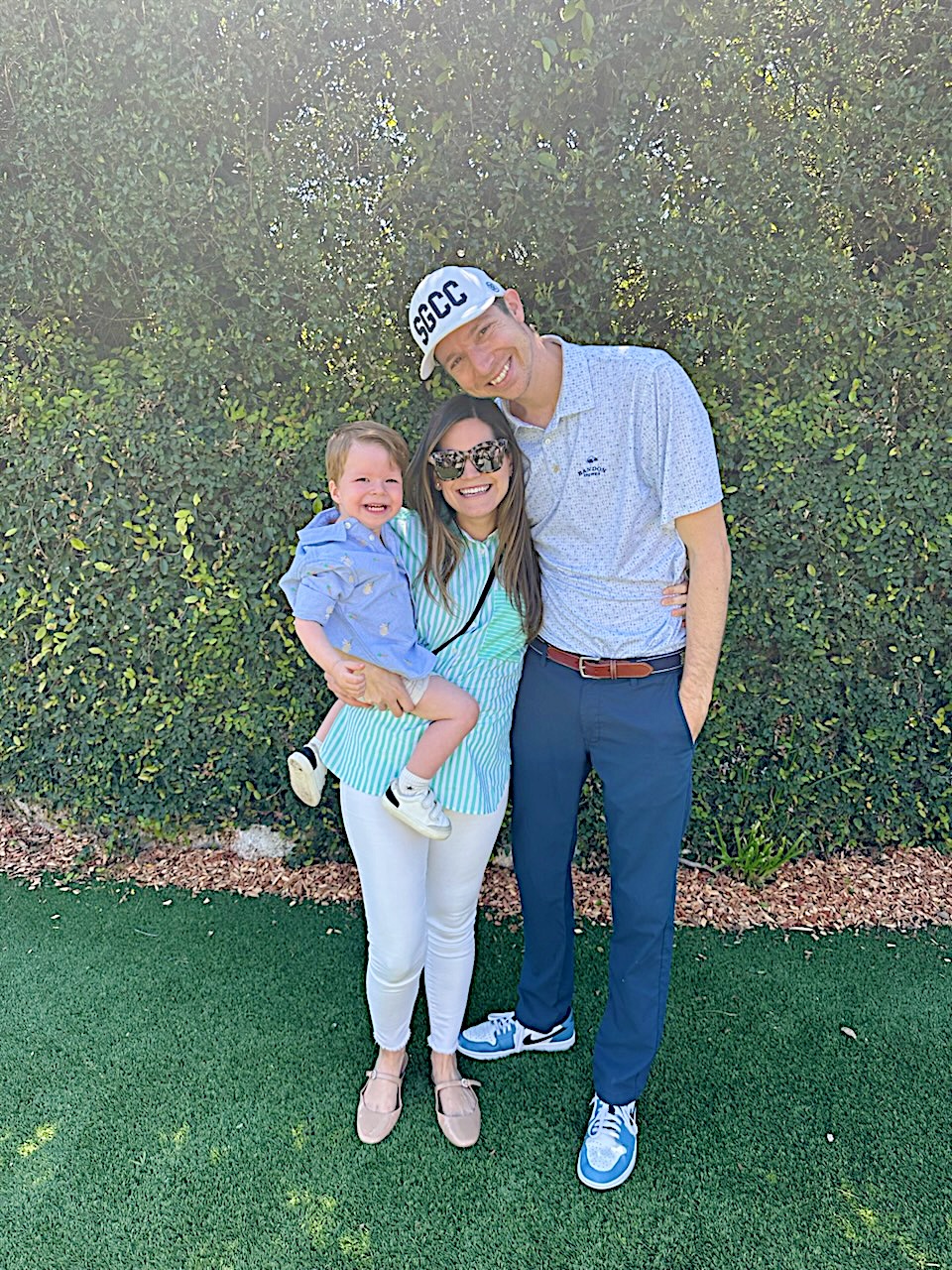 A man, woman, and young child stand together outdoors on green grass, smiling in front of a leafy hedge. The man wears a cap, and the woman holds the child in her arms.