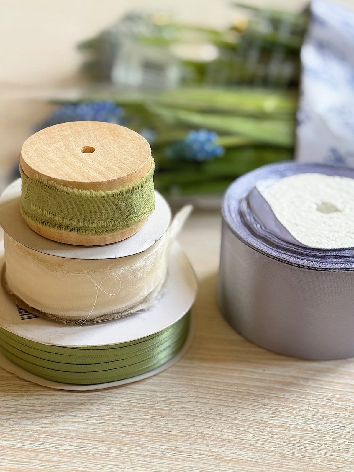 Spools of green, cream, and silver ribbon are stacked on a light wooden surface with blurred artificial flowers in the background.