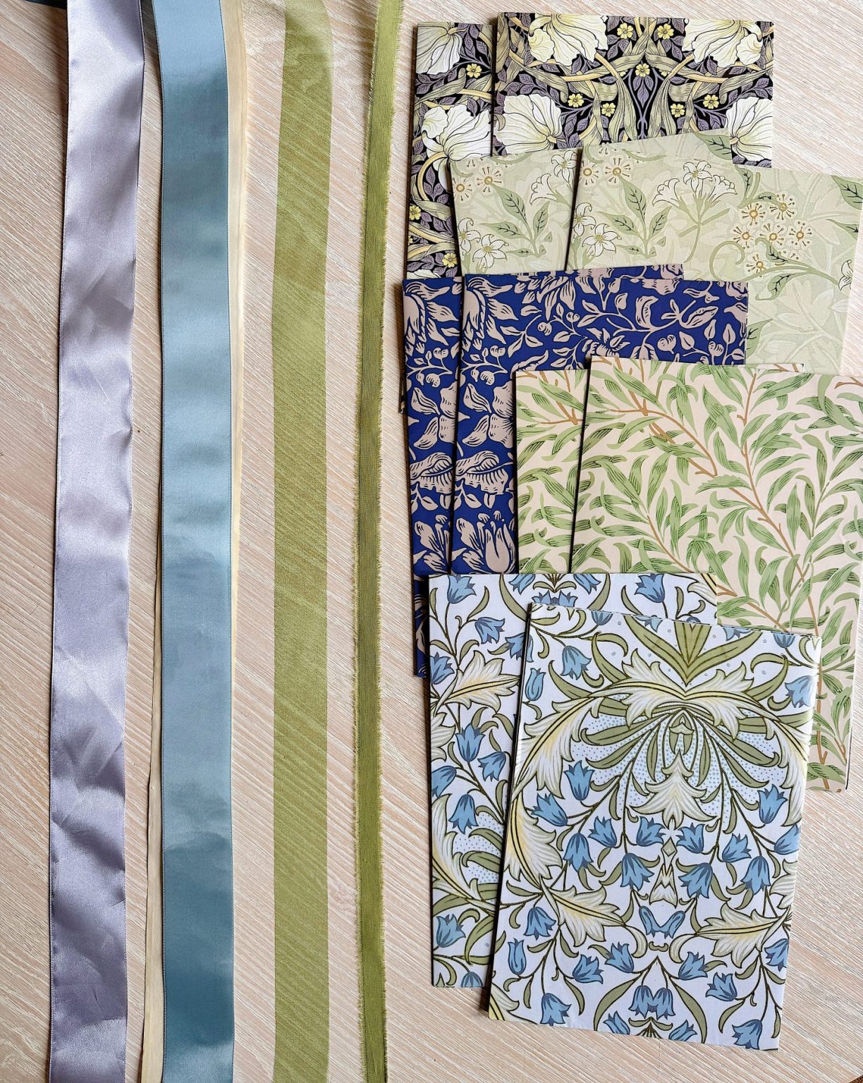 Four satin ribbons and six sheets of patterned paper with botanical designs are arranged side by side on a light wooden surface.