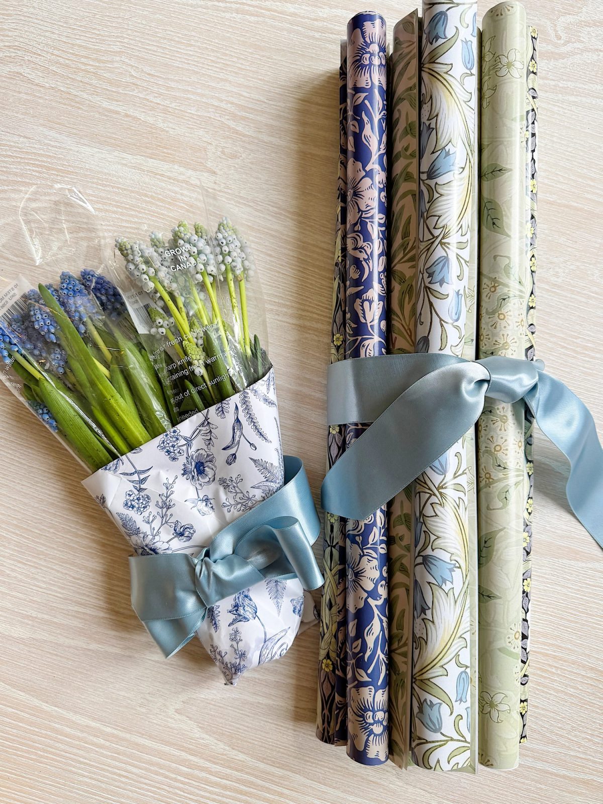 A bouquet of grape hyacinths wrapped in floral paper with a blue ribbon next to four rolls of floral-patterned wrapping paper tied with a blue ribbon on a light surface.