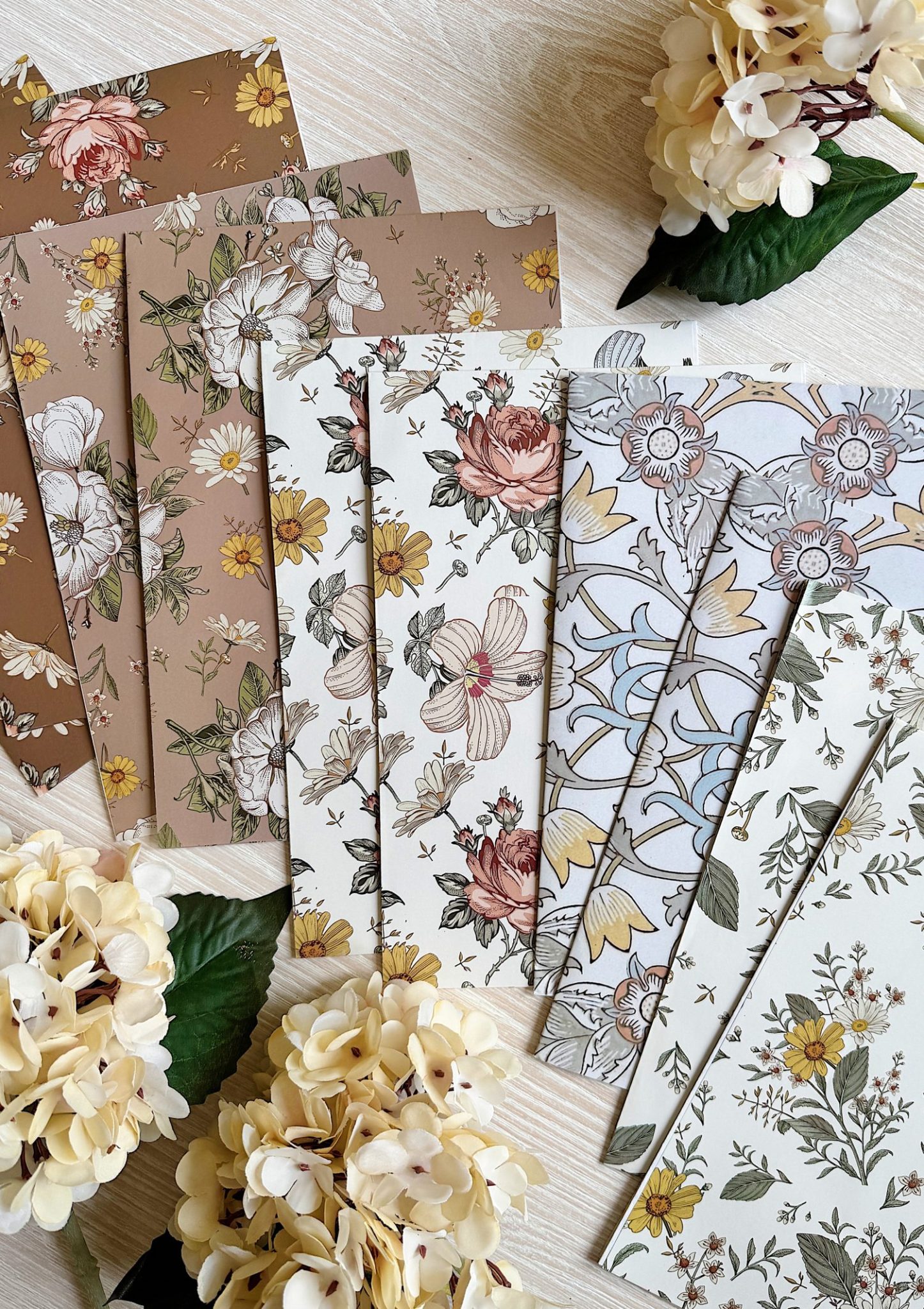 Several sheets of patterned paper featuring floral designs in brown, cream, and white tones are arranged on a light wood surface, surrounded by artificial cream-colored flowers.