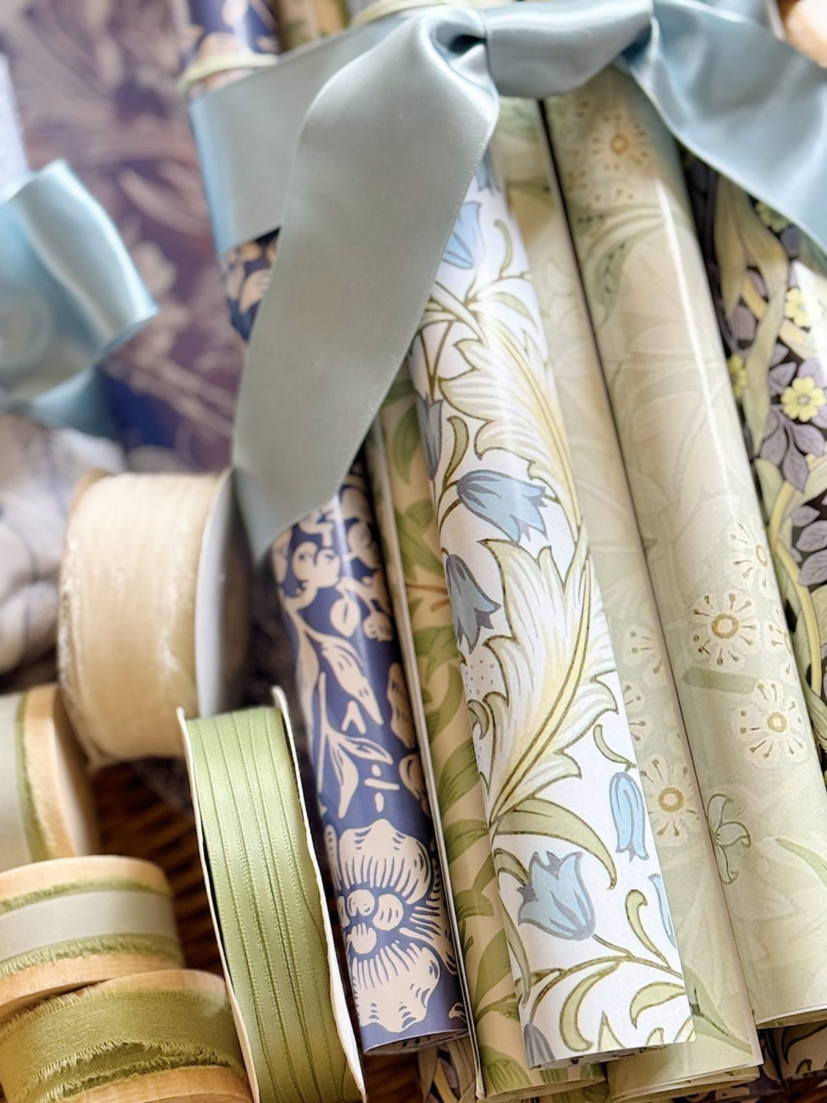 Rolled sheets of patterned wrapping paper in blue and green tones are tied with a blue ribbon, with matching colored spools of ribbon nearby.