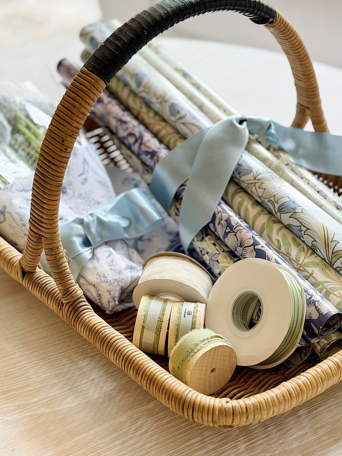 A wicker basket holding rolls of patterned wrapping paper, blue ribbon bows, and several spools of ribbon on a light wooden surface.