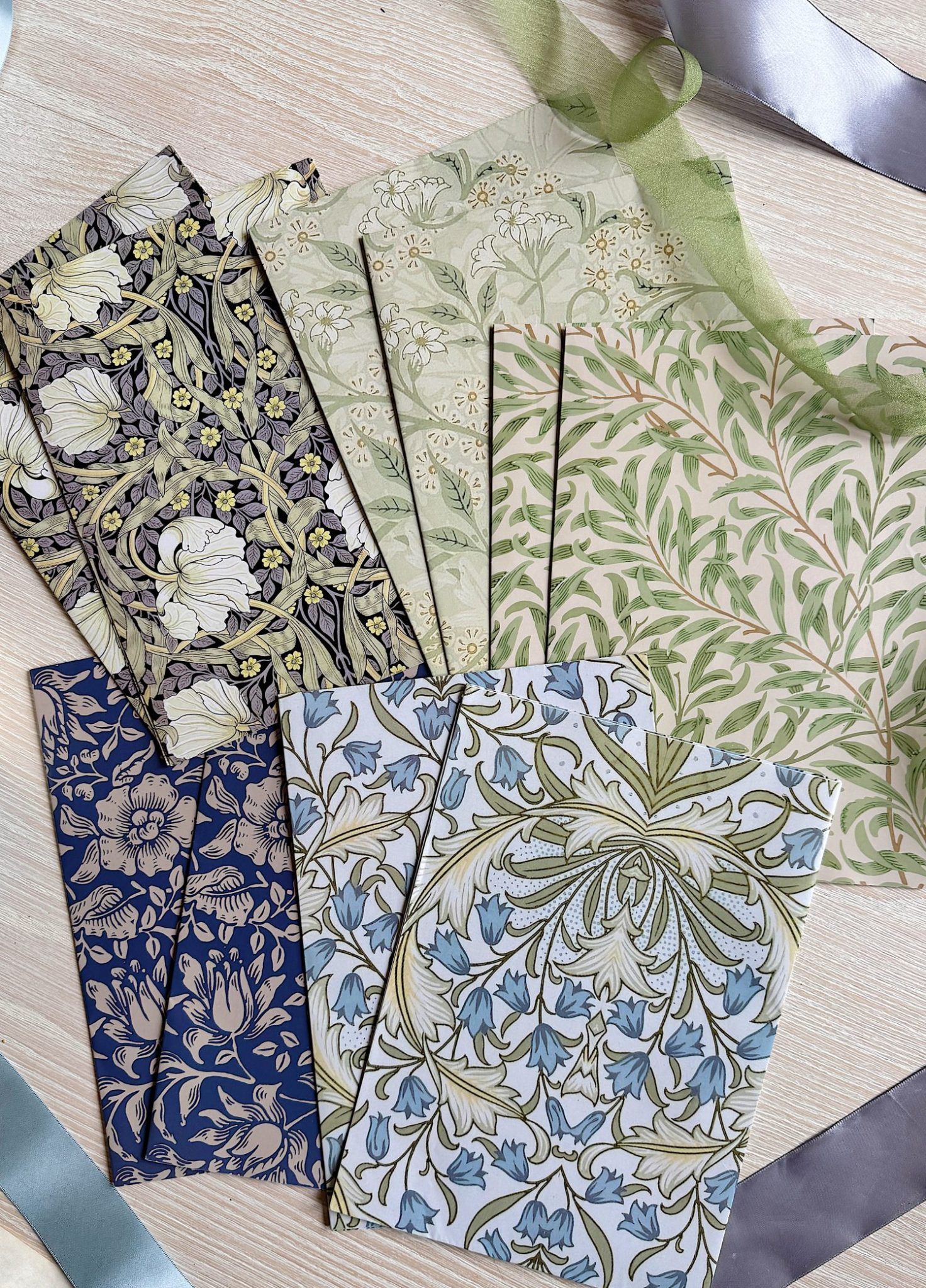 Six sets of patterned paper sheets with botanical and floral designs in green, blue, and beige tones are arranged on a light wooden surface, with satin ribbons partially visible around them.