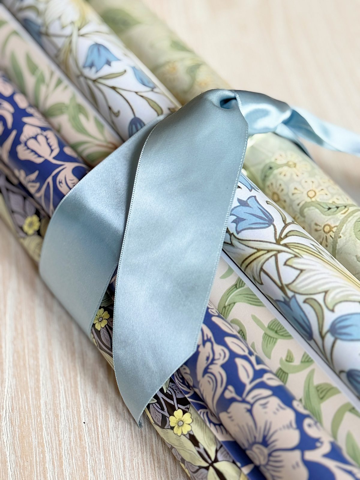 Three rolls of floral-patterned wrapping paper are tied together with a light blue satin ribbon on a light wooden surface.