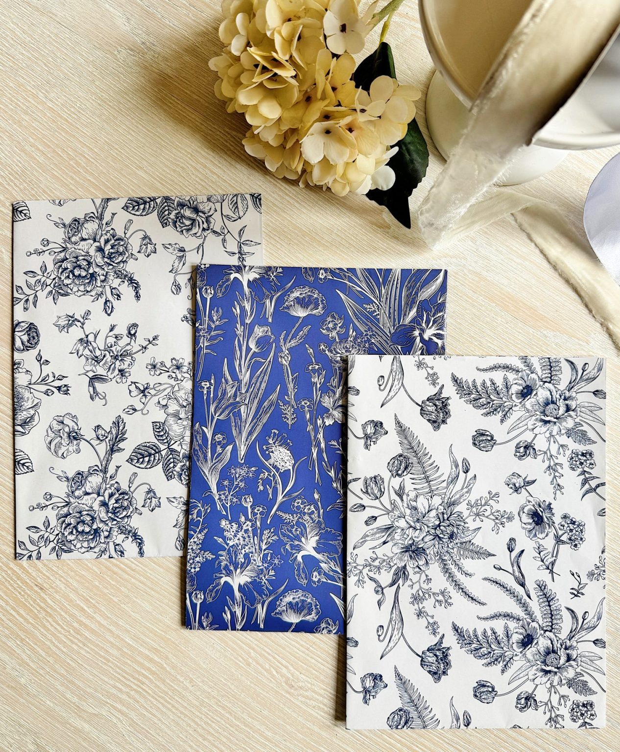 Three notebooks with floral-patterned covers, two in white with black designs and one in blue with white designs, are arranged on a light wooden surface next to a bouquet of pale flowers.
