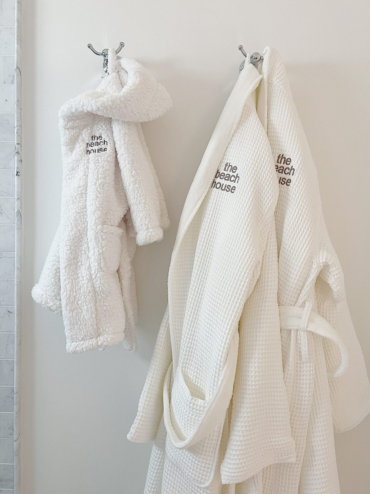 Two white bathrobes hang on hooks. One robe is fuzzy, and the other is waffle-textured. Both have "the beach house" embroidered on them.