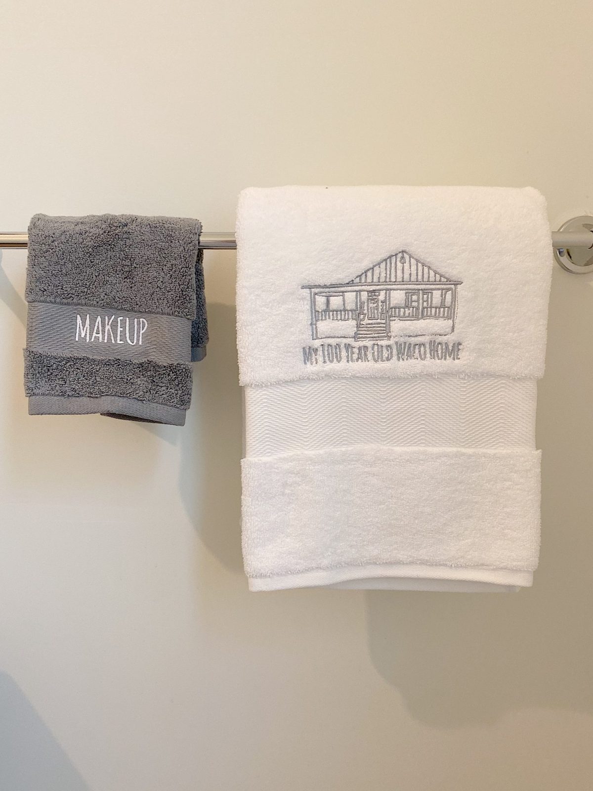 Two towels on a rod: a gray towel labeled "MAKEUP" on the left and a white towel with a house design and "MY 100 YEAR OLD WACO HOME" text on the right.