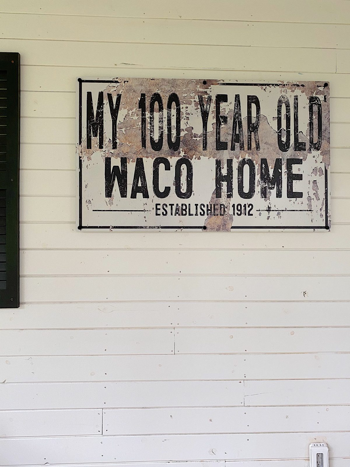 A sign on a light-colored wall reads, "My 100 Year Old Waco Home, Established 1912," with a weathered appearance.