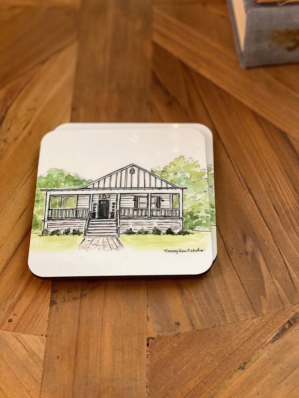 Illustrated coaster depicting a small, single-story house with a front porch and surrounding greenery, placed on a wooden table.