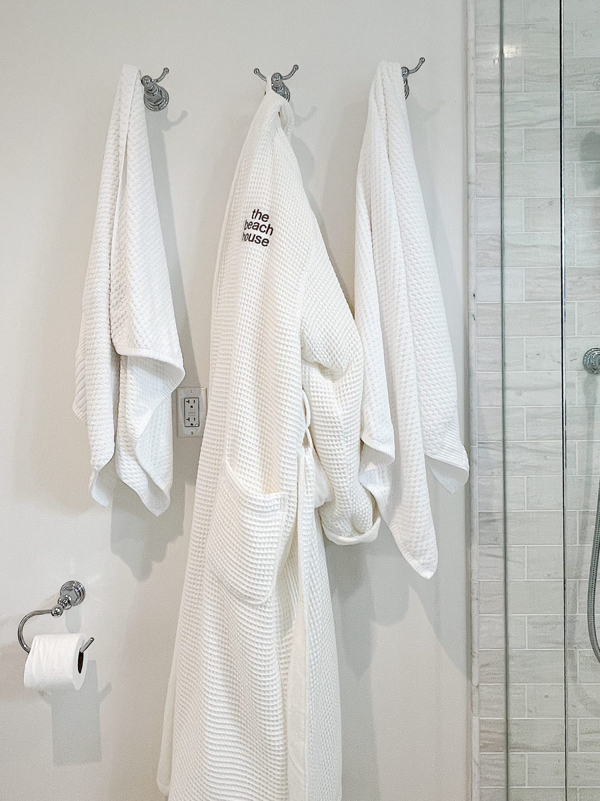 Three white towels hang on hooks beside a white shower wall. A bathrobe with "the beach house" text is in the center, next to a toilet paper holder.