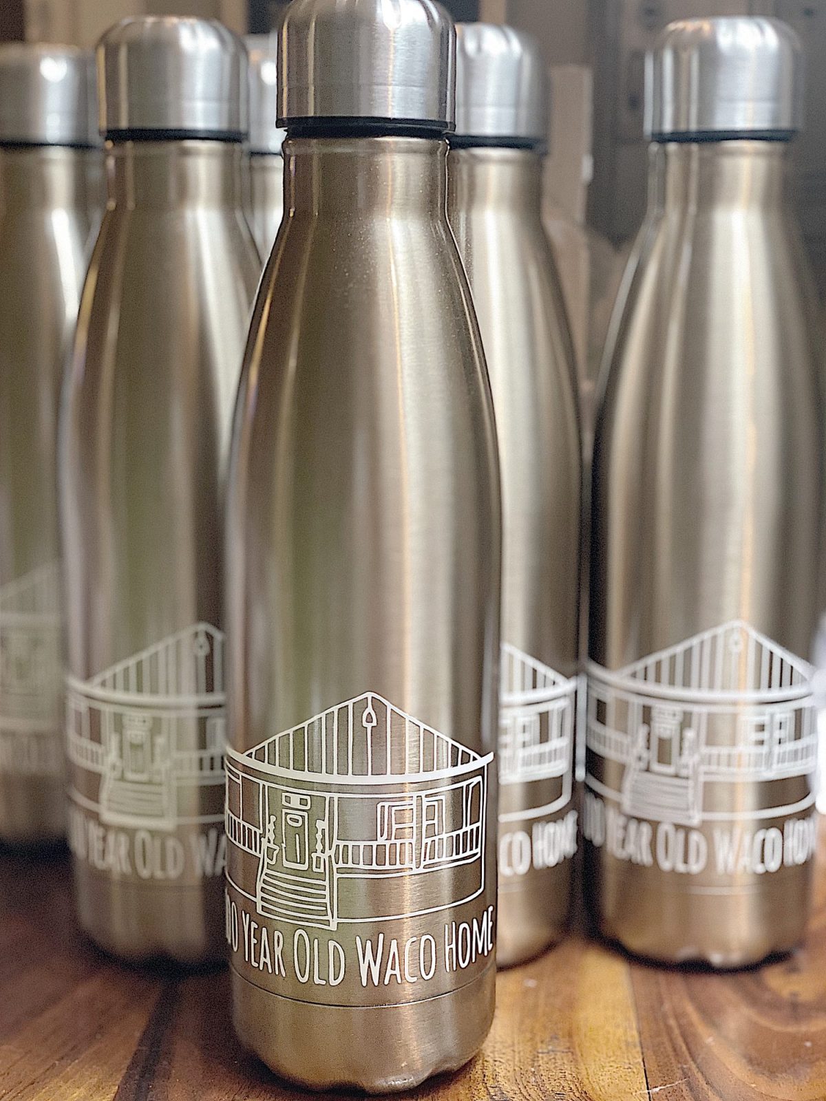 Metal water bottles with a house illustration and "110 Year Old Waco Home" text printed on them, arranged in rows.