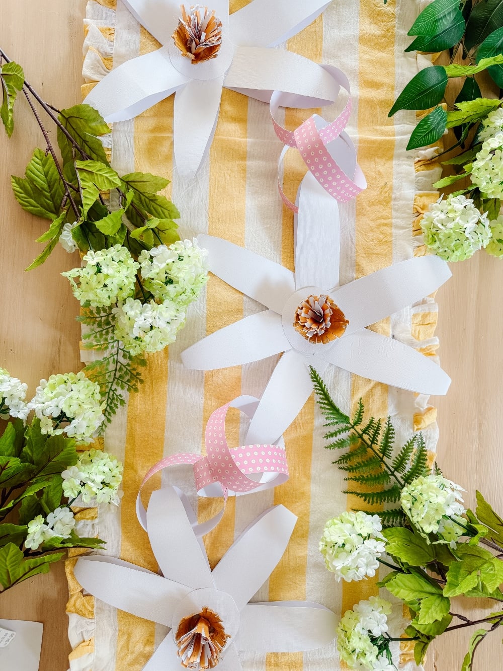 Paper flowers with pink and white ribbons are arranged on a yellow-striped table runner, surrounded by green leaves and white artificial flowers&mdash;perfect inspiration for your next paper craft ideas.