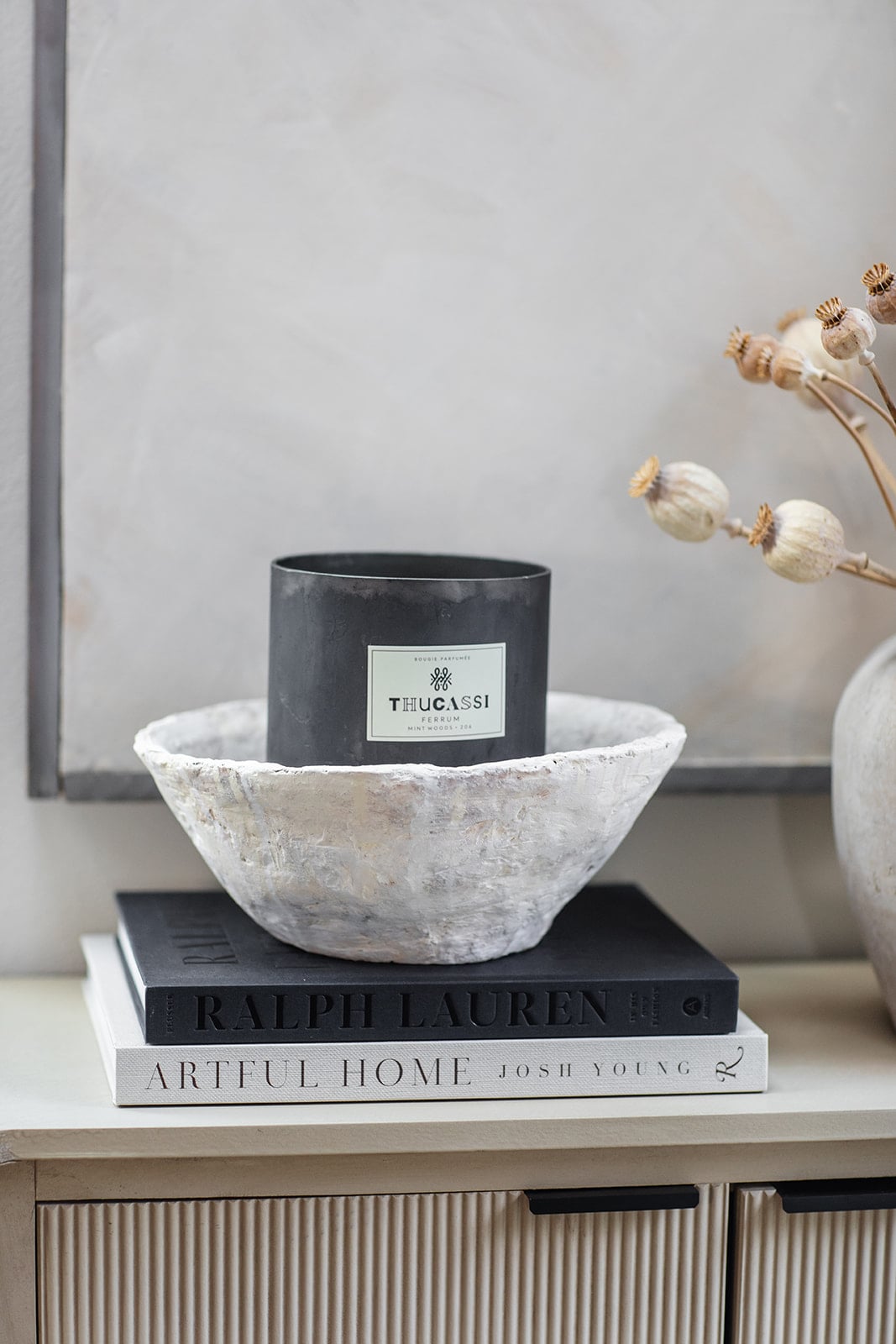 A black Thucassi candle in a white bowl sits on two stacked books, "RALPH LAUREN" and "ARTFUL HOME," on a light-colored surface next to dried plants&mdash;adding inspiration for paper craft ideas to your decor.