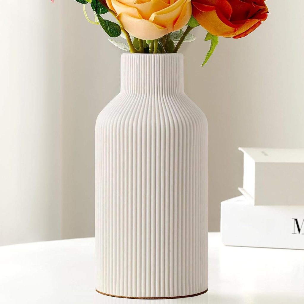 A white ribbed ceramic vase holding yellow and orange artificial roses, inspired by summer wedding flowers, sits on a white surface next to a stack of books.