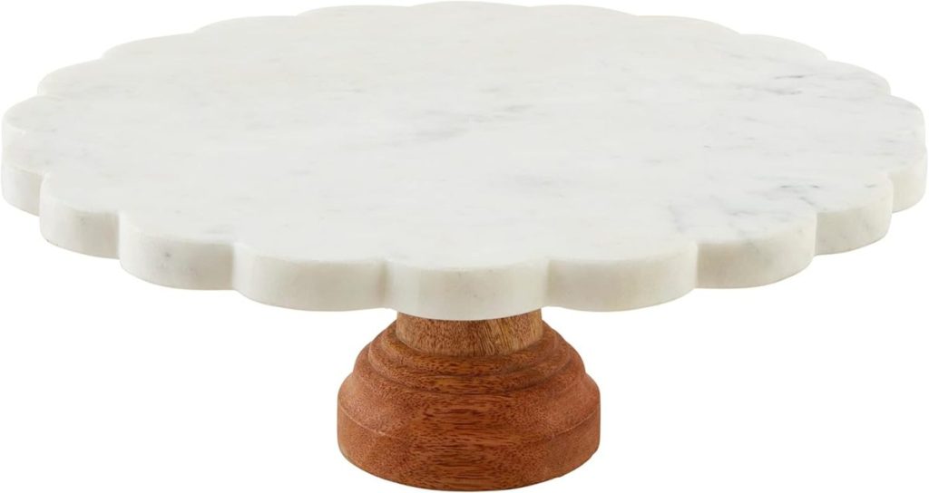 A round, white marble cake stand with a scalloped edge sits atop a short, brown wooden base—perfect for displaying your favorite spring cake.