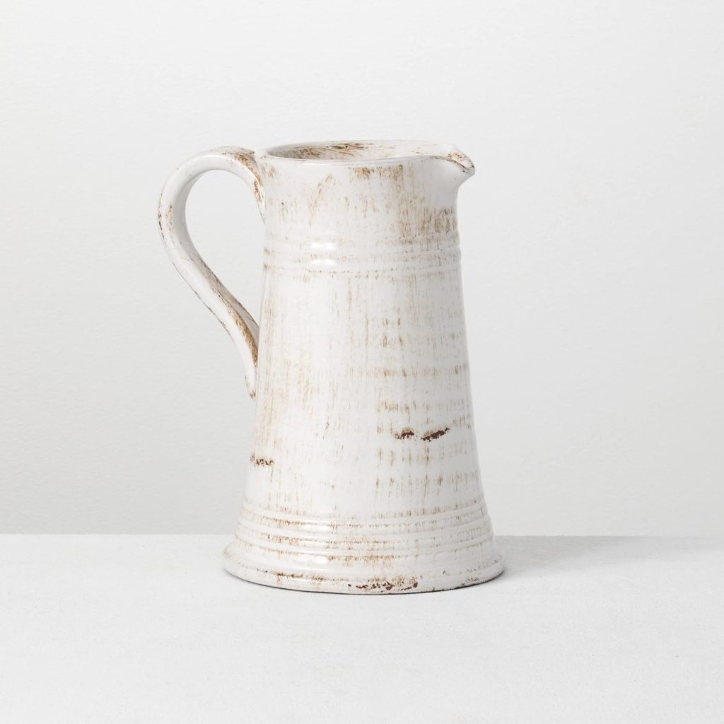 A white ceramic pitcher with a handle and spout, featuring a distressed, rustic finish, brings farmhouse kitchen charm as it sits on a plain white surface against a white background.