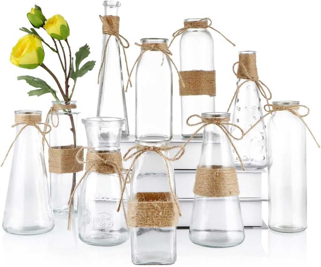A collection of ten clear glass vases, each decorated with twine, with two yellow roses in one vase&mdash;perfect summer wedding flowers&mdash;arranged on and in front of white books.