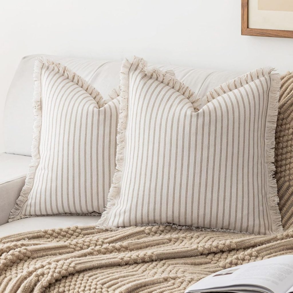 Two cream-colored pillows with thin brown stripes and fringe edges rest on a beige textured blanket, placed on a light-colored sofa—perfect for effortless summer decorating ideas.