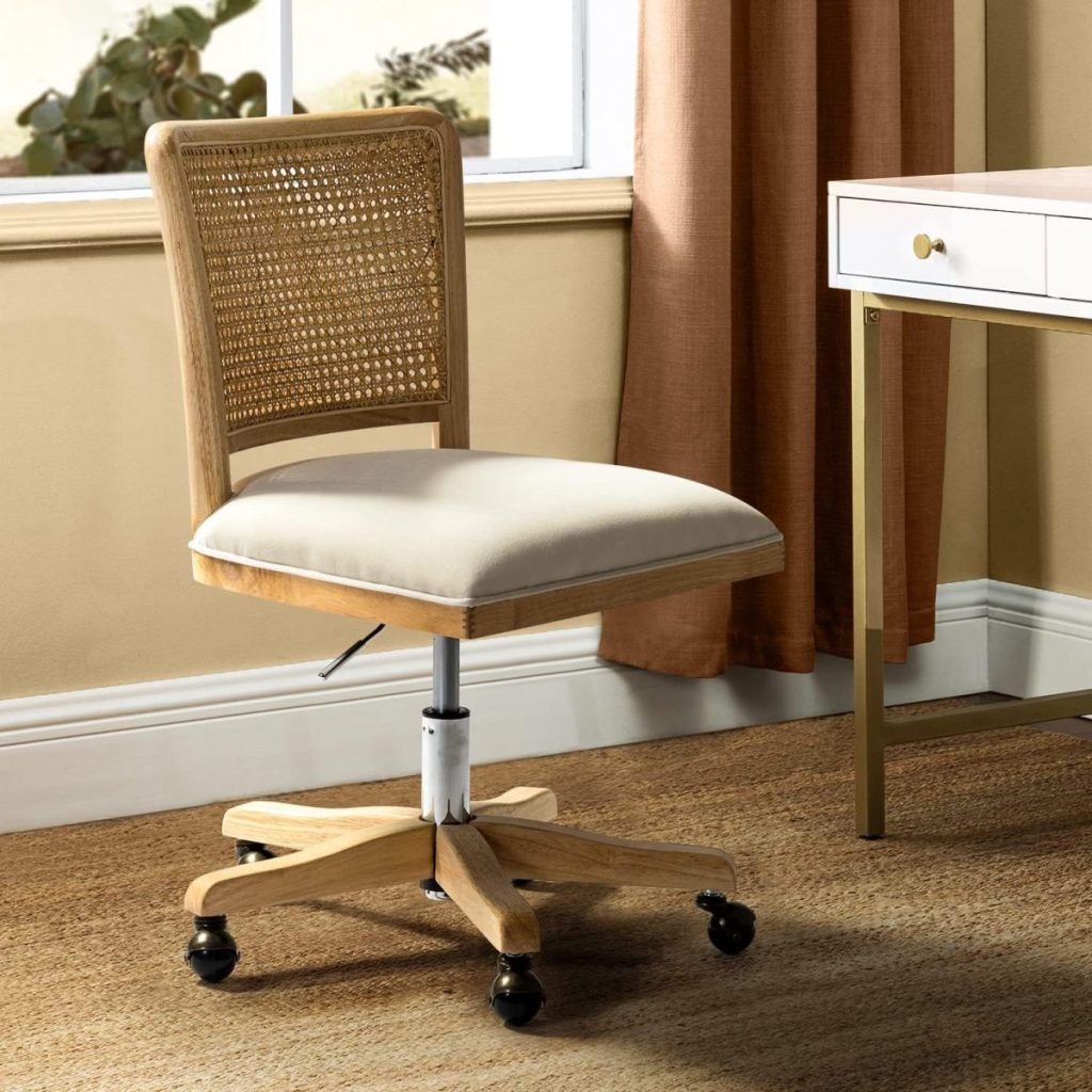 A wooden office chair with a cane backrest, beige cushioned seat, and caster wheels sits next to a desk in a sunlit room overlooking an open concept kitchen.