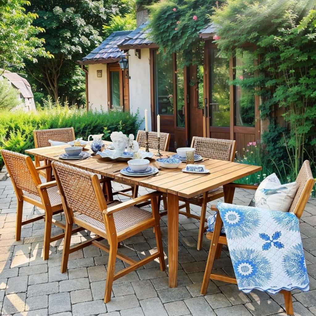 A wooden outdoor dining table set with dishes and six chairs, some accented with outdoor cushions, sits on a stone patio near a cottage, surrounded by greenery and a blue-patterned blanket draped over one chair.