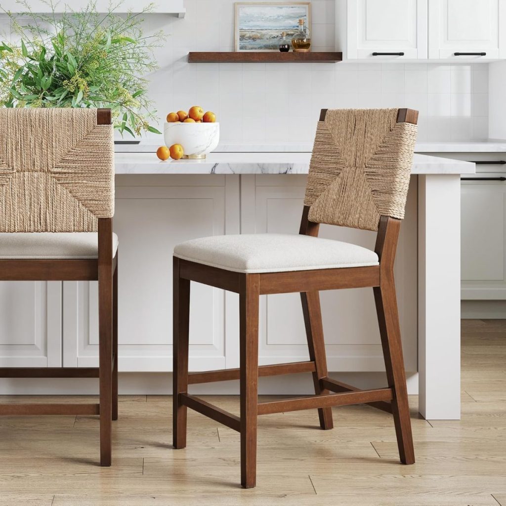 A wooden bar stool with a woven backrest and cushioned seat sits in a bright farmhouse kitchen with white cabinets and a bowl of oranges on the counter.