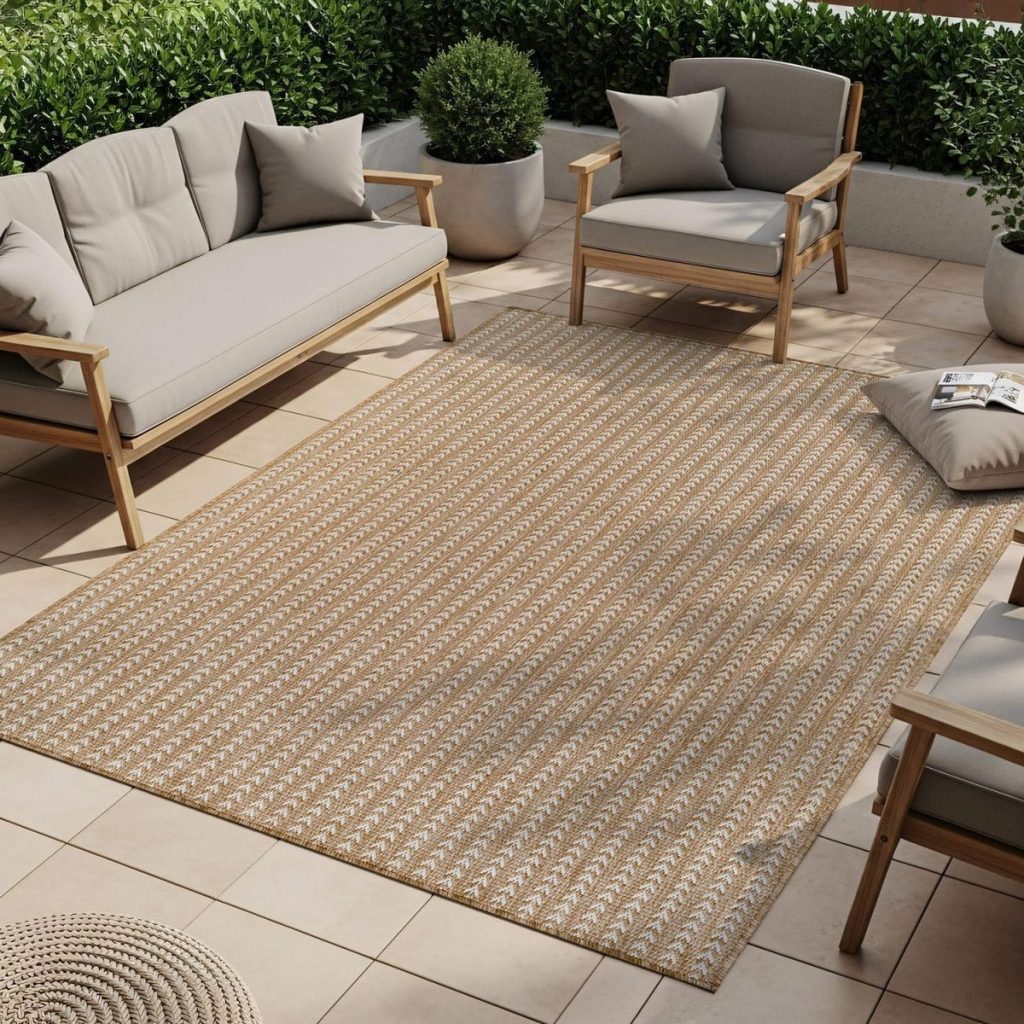Outdoor patio setup with light wooden furniture, gray cushions, and a patterned beige area rug on beige tiles; potted plants line the background—perfect for inspiring your Summer Decorating Ideas.
