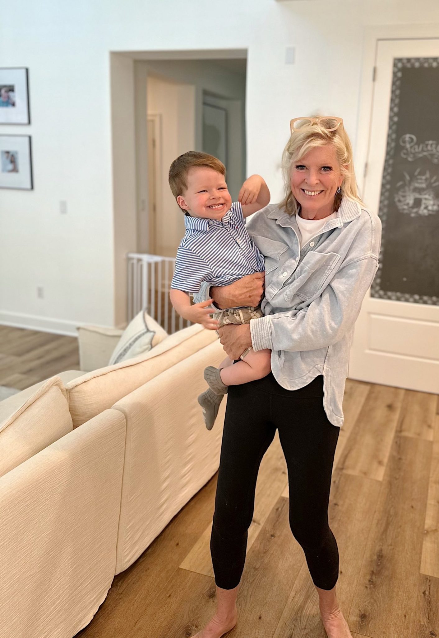 A woman stands indoors, smiling and holding a young boy who is also smiling. They are in a living room with a white couch and wooden floor.