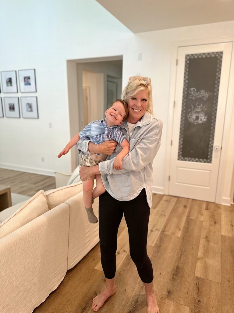 A woman stands barefoot on a wooden floor, smiling and holding a laughing young child in a casual, well-lit living room.
