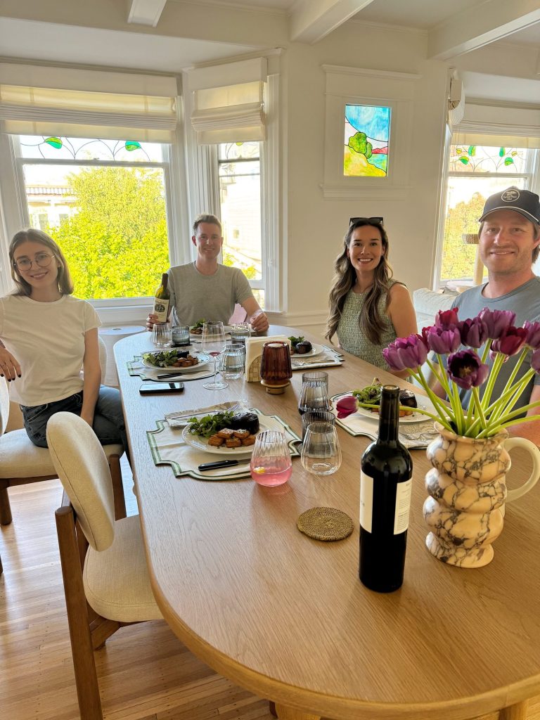 Four people sit around a dining table set with food and drinks in a bright room with large windows and purple tulips in a vase.