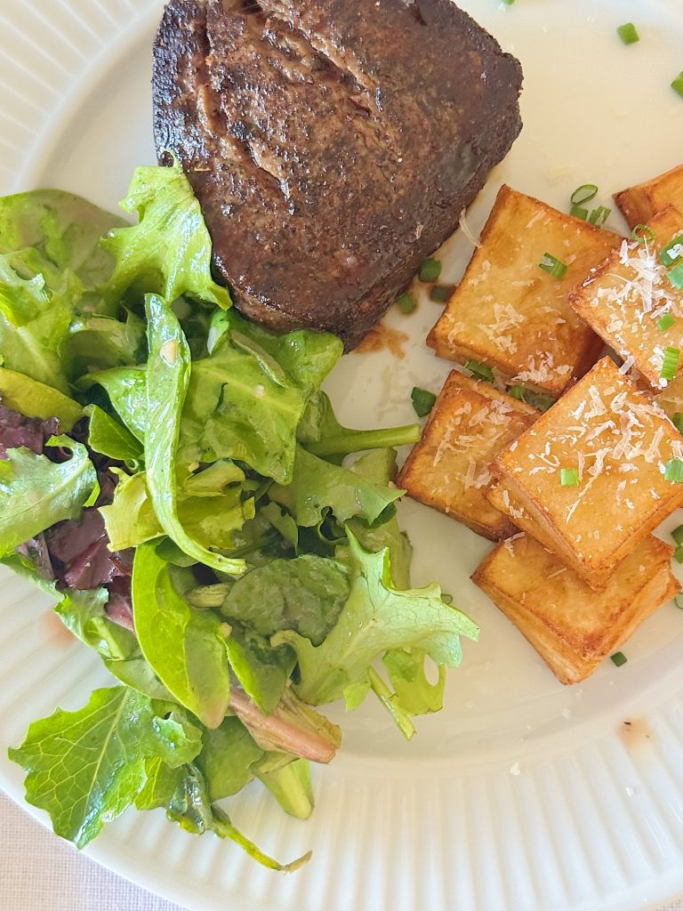 A plate with a piece of grilled steak, a serving of green salad, and several golden brown roasted potato cubes topped with grated cheese and chopped chives.