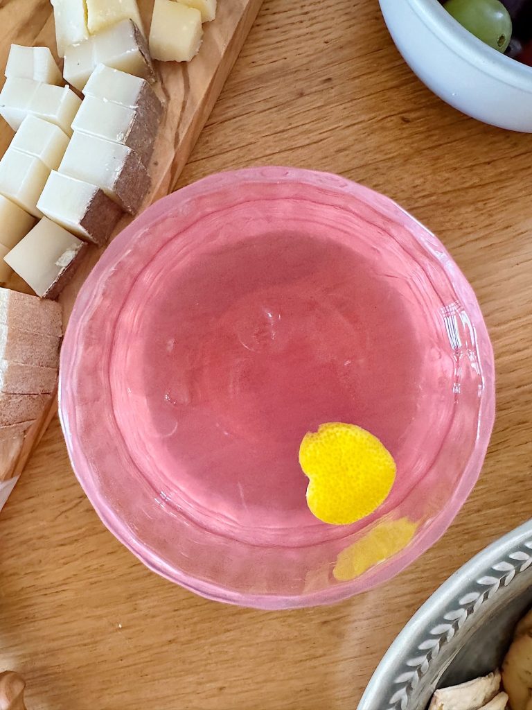 A pink cocktail in a glass with a lemon peel garnish on a wooden table, surrounded by cheese cubes and bowls.