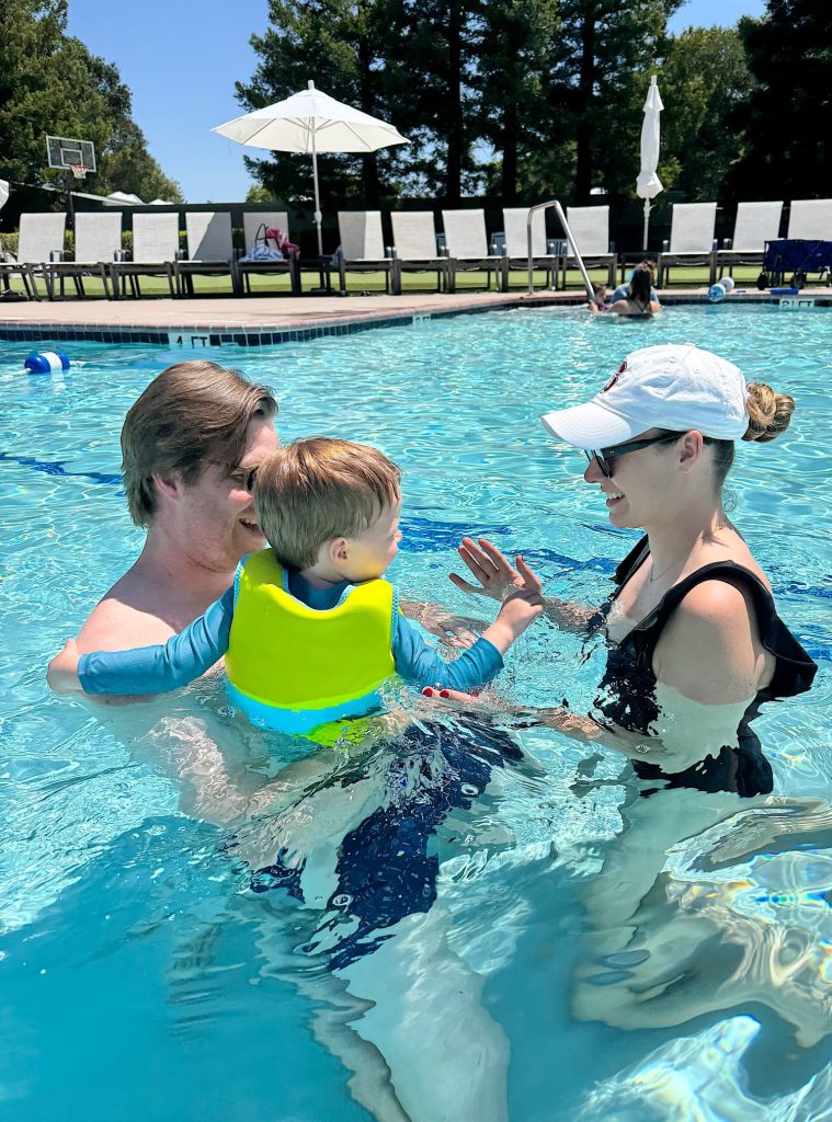 A man and woman in a swimming pool help a young child wearing a yellow life vest; lounge chairs and umbrellas are visible in the background.