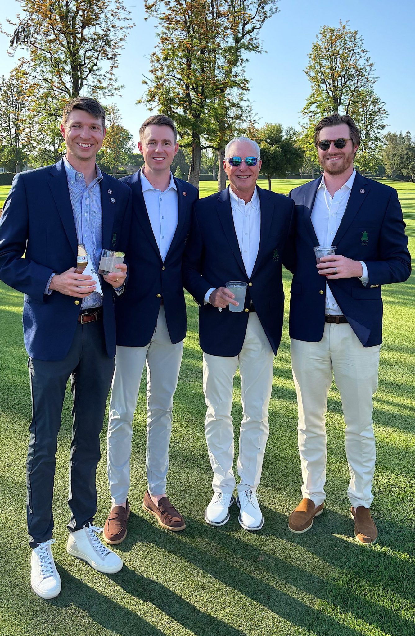 Four men in blue blazers and light pants stand on a golf course, smiling and holding drinks on a sunny day.