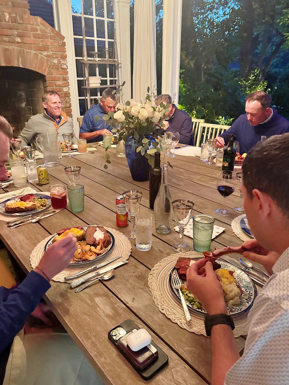 Six men sit around a wooden dining table eating dinner, with plates of food, drinks, and a flower centerpiece in the middle. Evening light filters through large windows in the background.