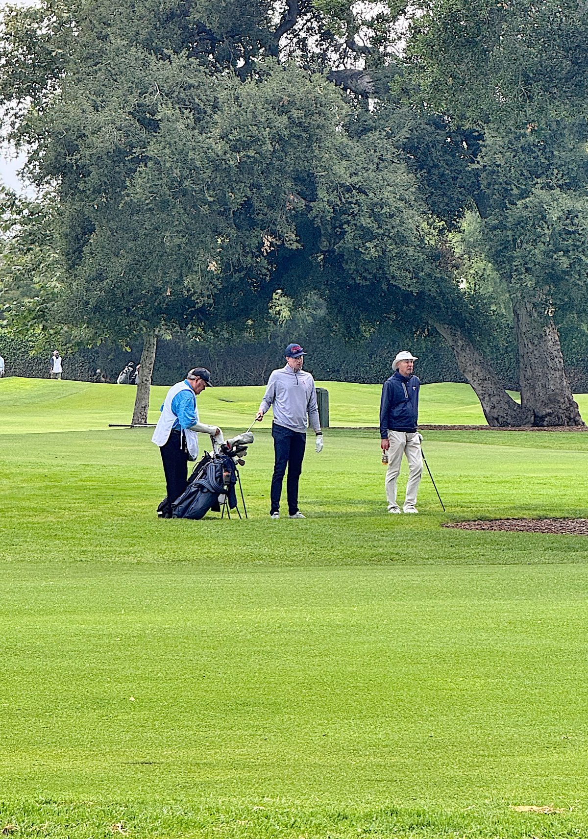 Three people stand on a golf course; one holds a golf bag, another has a golf glove, and the third holds a club, with trees and greenery in the background.