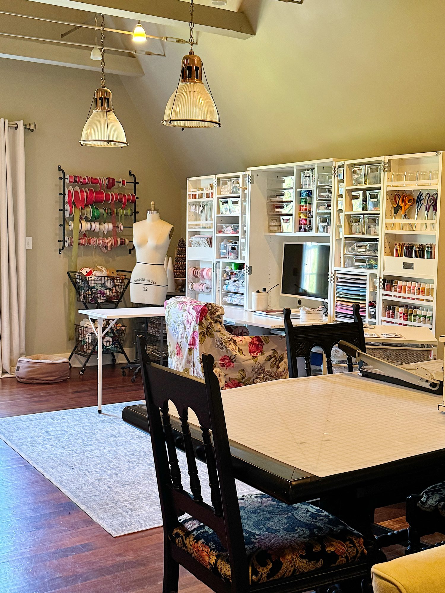 A craft room with organized shelves of supplies, a table, chairs, a mannequin, a computer monitor, and a rack of colorful ribbons.