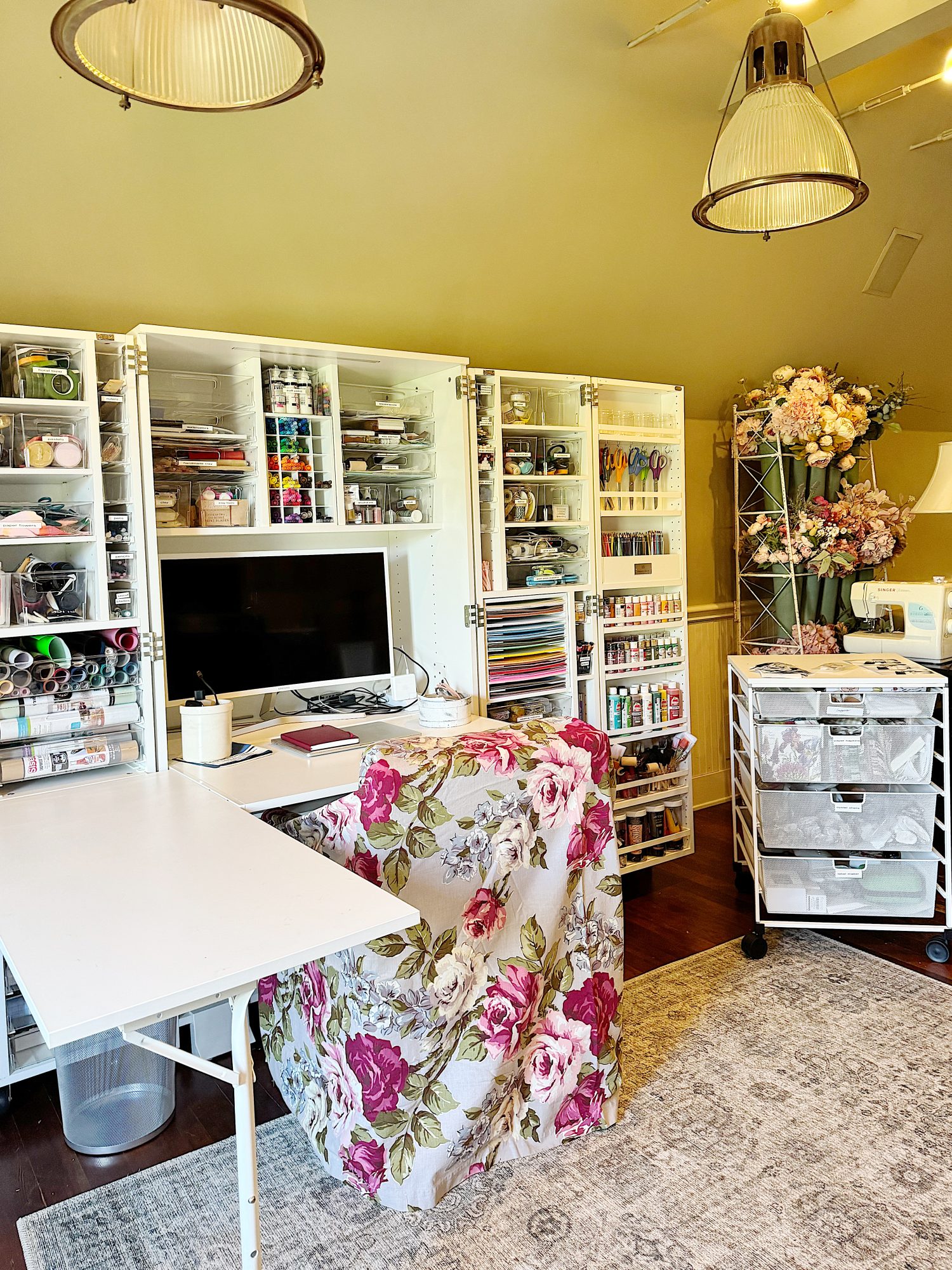 A craft room with organized shelves of supplies, a computer monitor, a desk covered with a floral cloth, storage carts, and artificial flowers in the corner.
