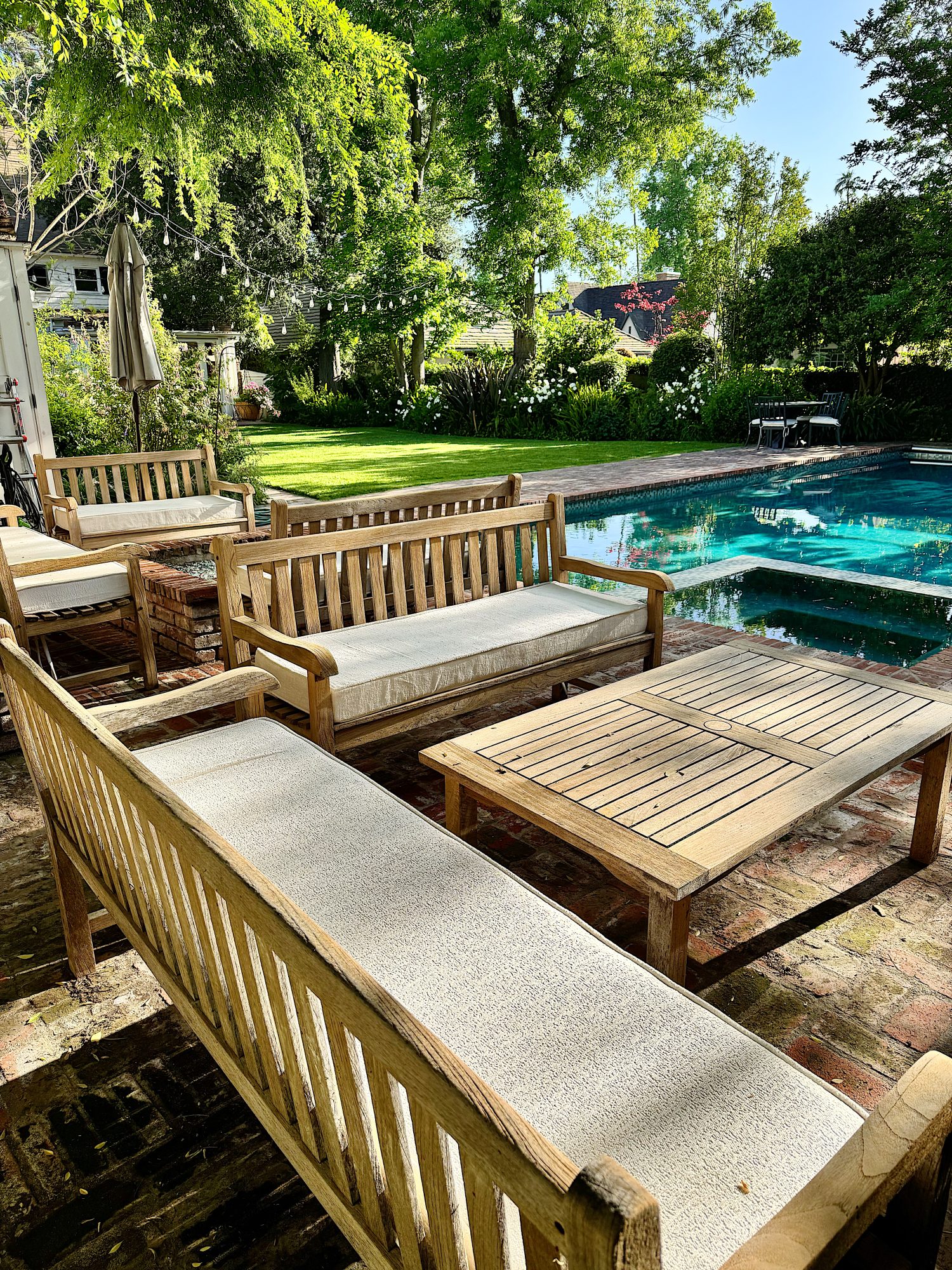 Wooden outdoor benches and tables with light cushions are arranged by a swimming pool in a backyard, surrounded by green trees and sunlight.