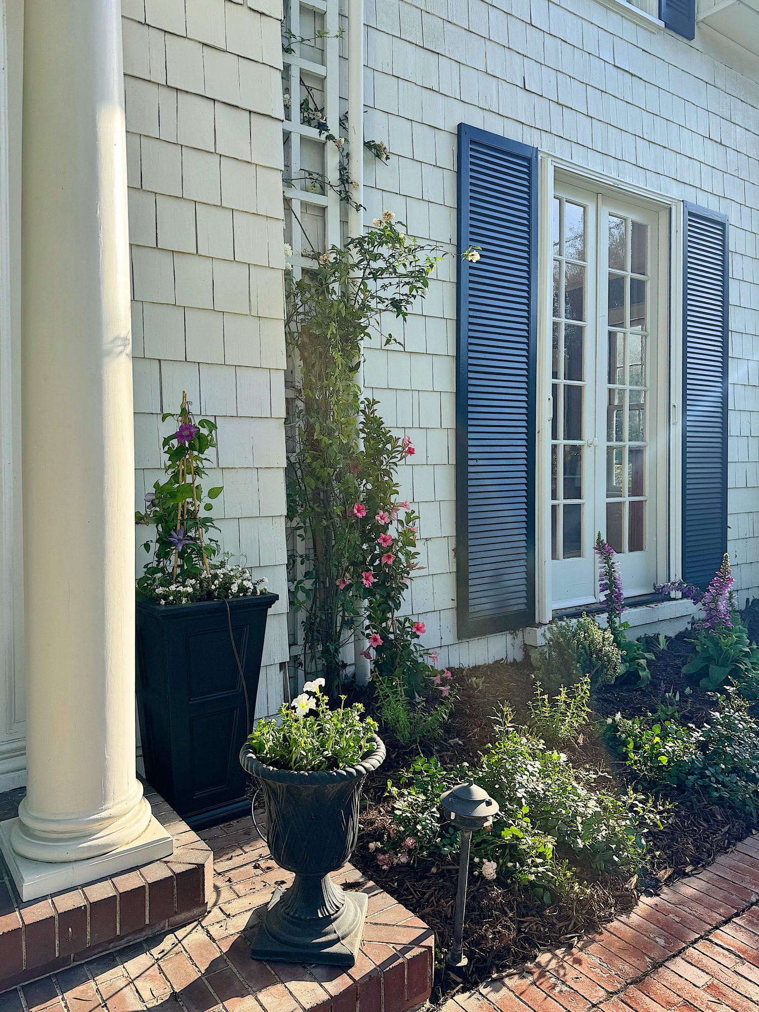 A white-paneled house features blue shutters, a glass door, potted plants, a small landscaped garden bed, and a brick walkway in sunlight.