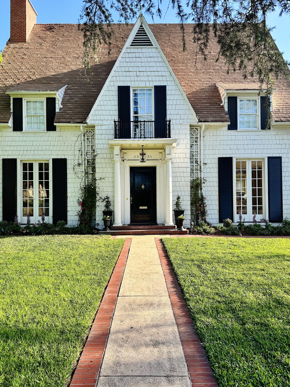 A white two-story house with dark shutters, a steep roof, and a brick chimney, viewed from the front with a straight walkway leading to the entrance.