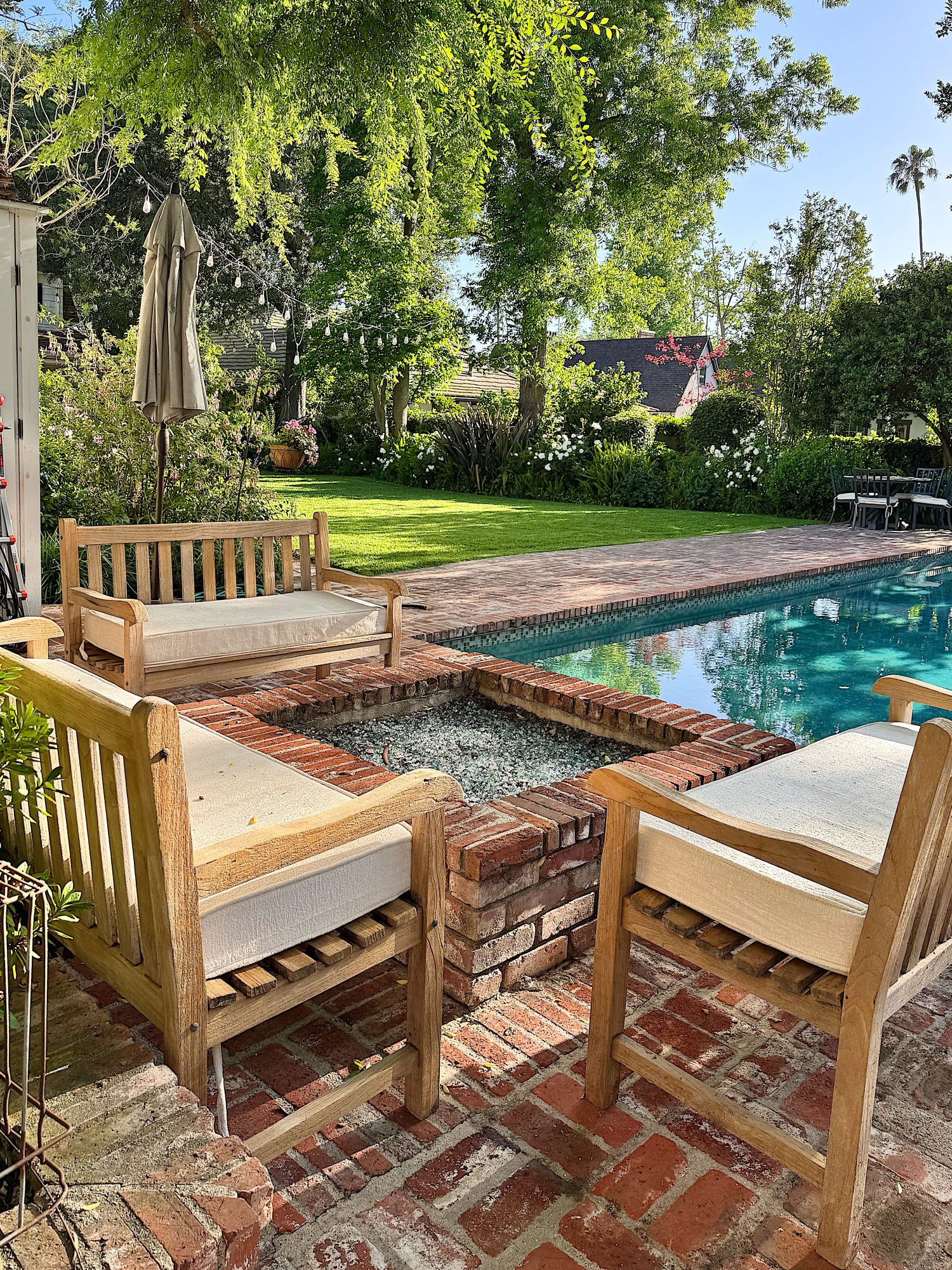 Wooden patio chairs surround a brick fire pit beside a swimming pool in a lush backyard with green lawn and trees.