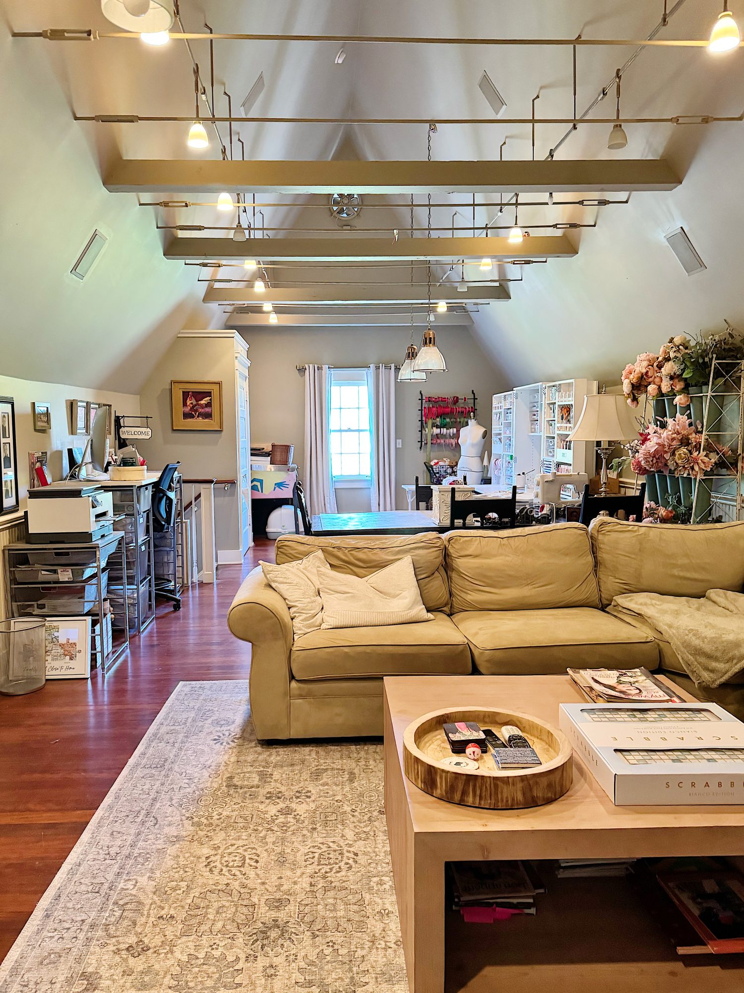 A spacious attic room with a beige sofa, coffee table, workstations, shelves, and craft supplies, featuring hardwood floors and natural light from a large window.