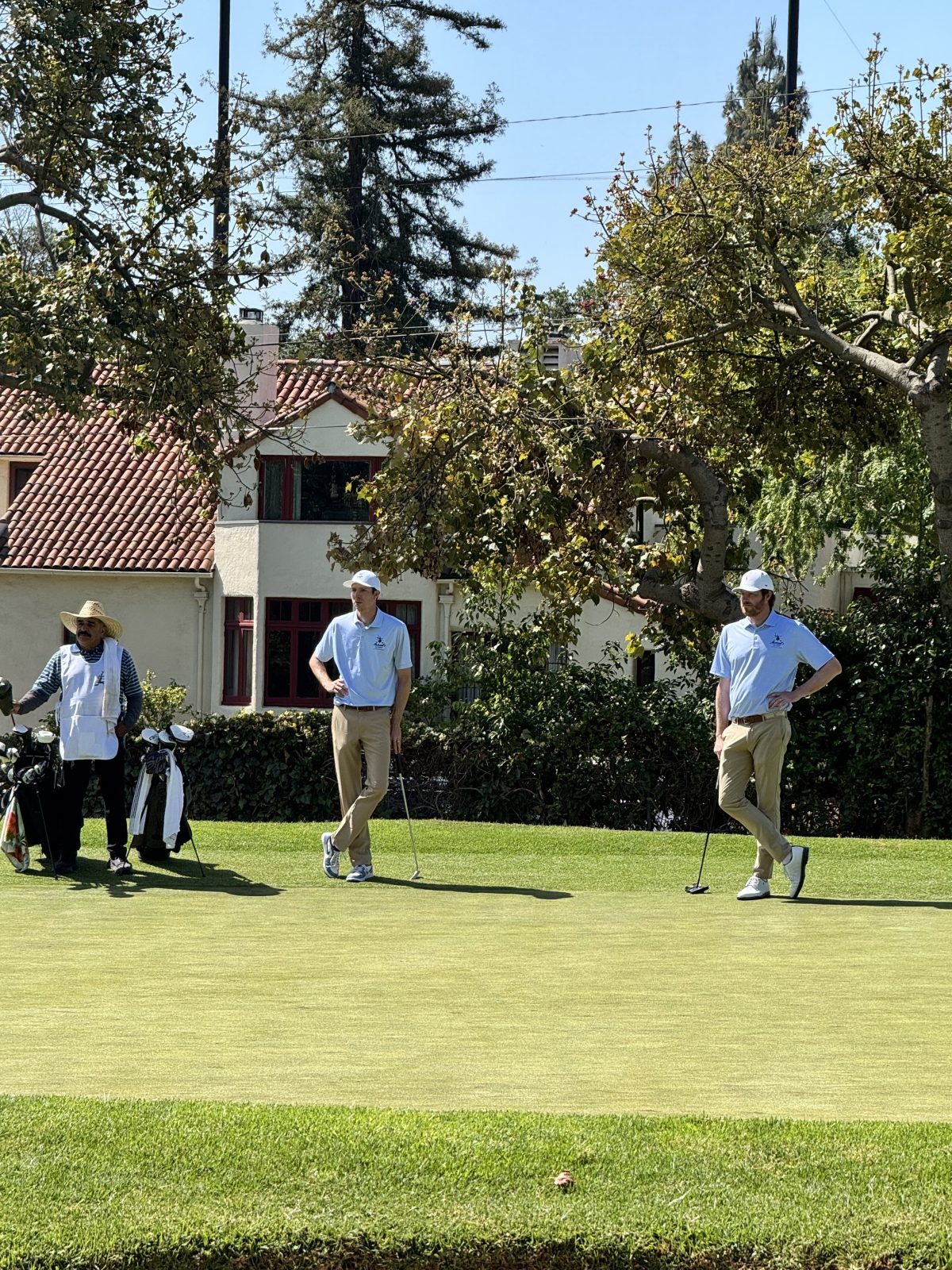 Two golfers in light blue shirts and khaki pants stand on a green near a caddie, with trees and a building in the background on a sunny day.