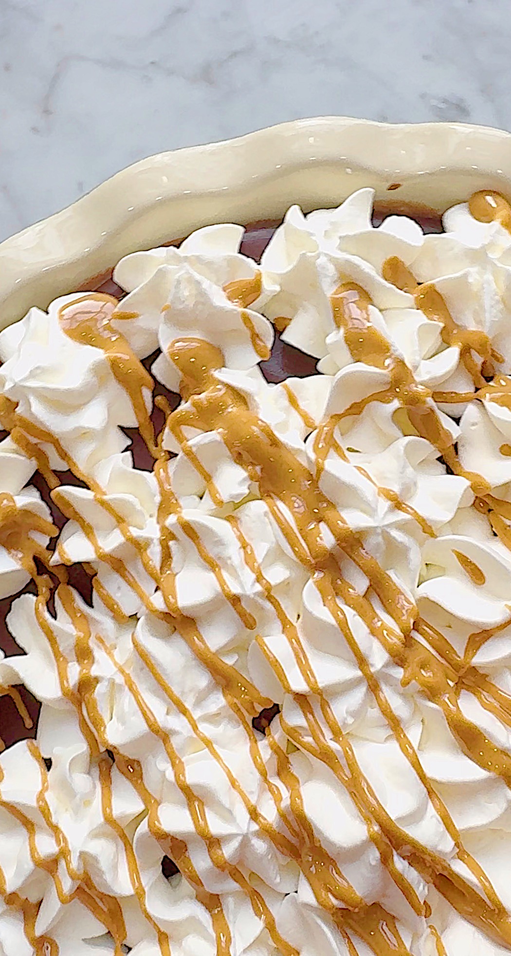 A pie topped with swirls of whipped cream and drizzled with caramel sauce, shown in a close-up view.